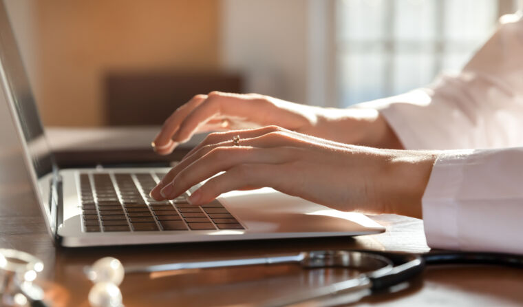 Hands of GP typing on laptop next to stethoscope