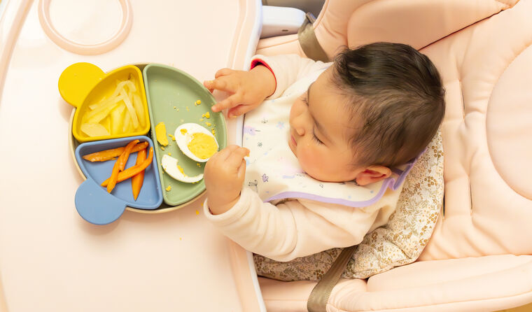 A baby sits at high chair with food, including egg