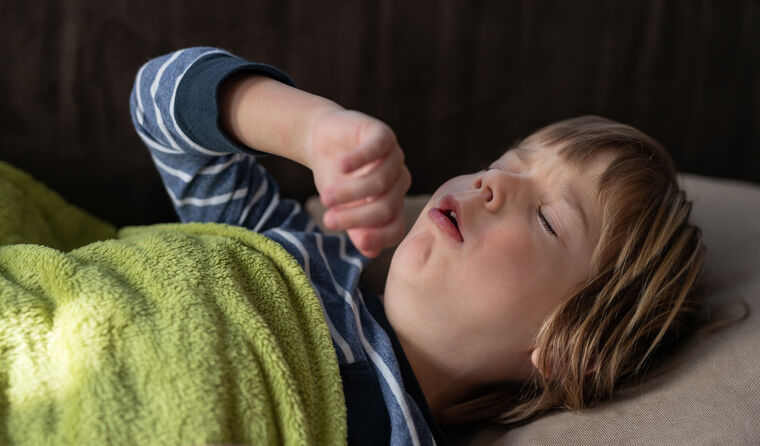 A young boy lies in bed coughing.